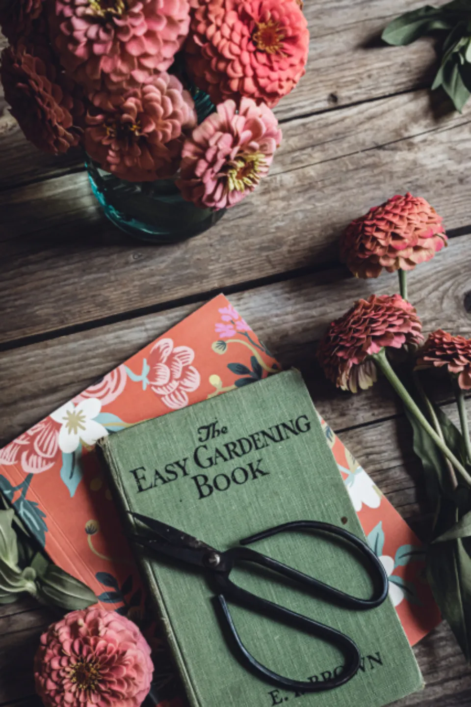Zinnias in a glass jar, an old gardening book and florist scissors on a wooden table