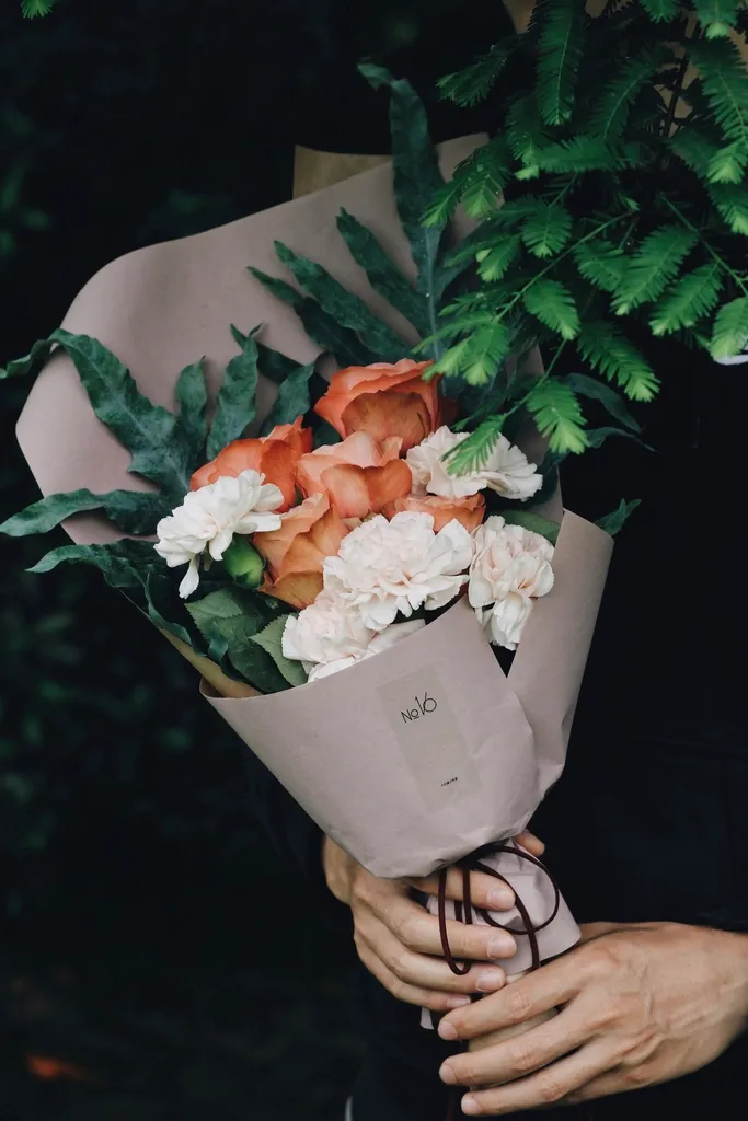 Hands holding a bouquet of peach roses and white carnations wrapped in kraft paper